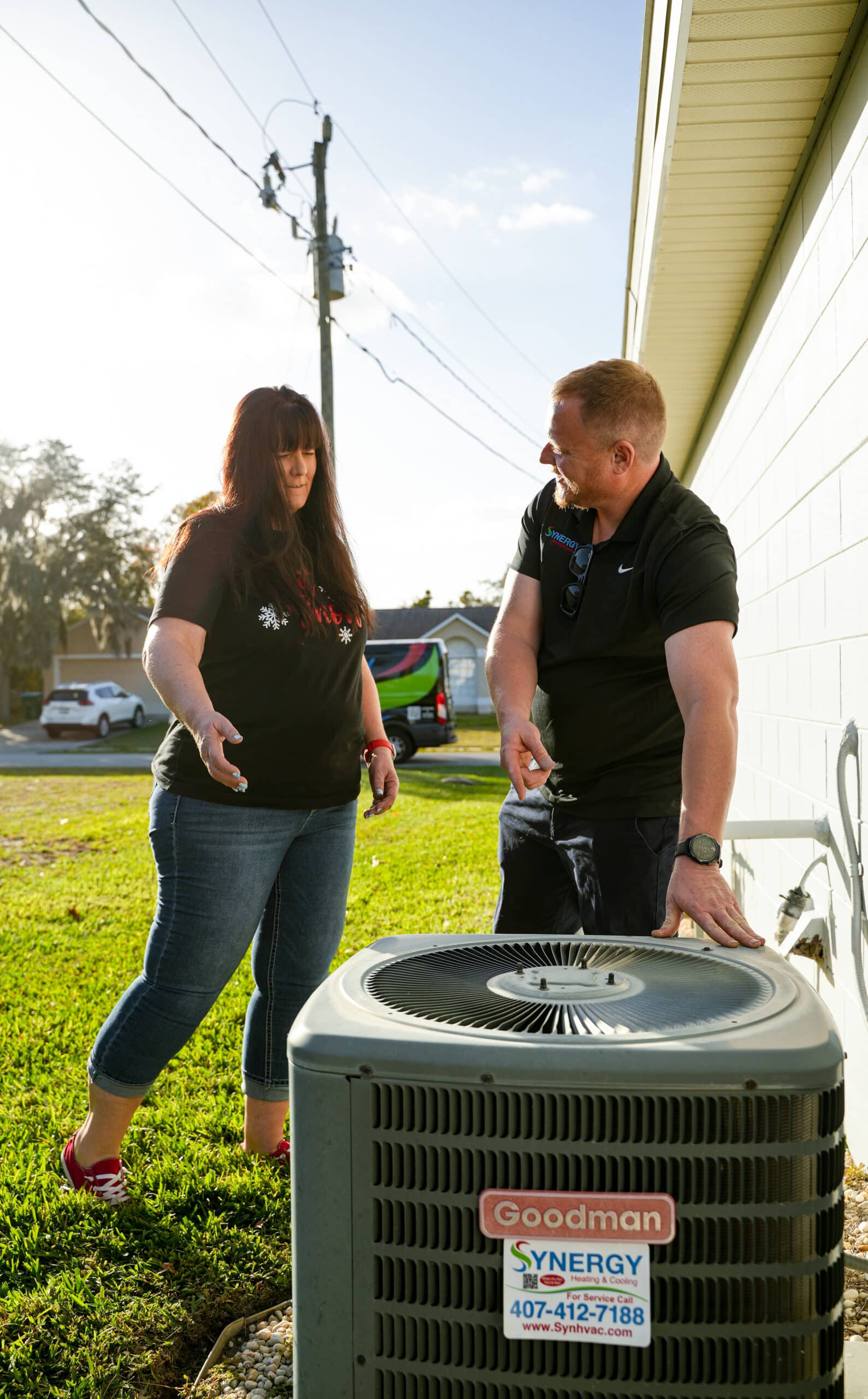 A Synergy Heating & Cooling technician showing a homeowner an outdoor AC condenser unit